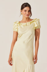 A woman is wearing an elegant cream-colored satin dress with a round neckline and short, puffed sleeves. The dress features ruffled details along the neckline. She has dark brown hair styled in a loose updo, and is accessorized with gold earrings.